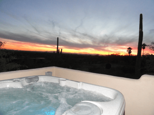 A hot tub with bubbling water overlooks a vibrant sunset and desert landscape with cacti and palm trees.