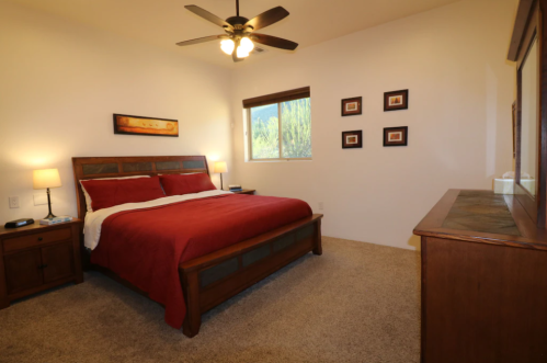 Cozy bedroom with a red bedspread, wooden furniture, and a window overlooking greenery. Soft lighting from lamps.