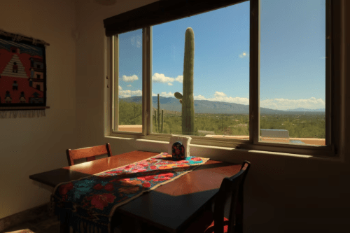 A cozy dining area with a colorful tablecloth, featuring a large window showcasing a desert landscape and a cactus outside.