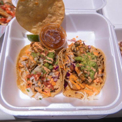 Two tacos filled with grilled meat, topped with slaw and sauces, served with a lime wedge and a small container of salsa.
