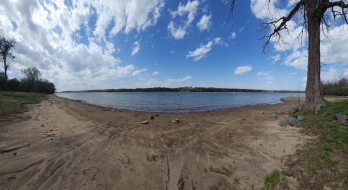 A panoramic view of a sandy beach by a calm lake, with blue skies and scattered clouds in the background.