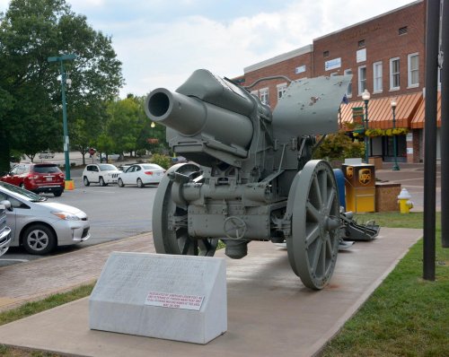 A historic cannon displayed on a concrete pedestal, with a plaque nearby, set in a town square with cars in the background.