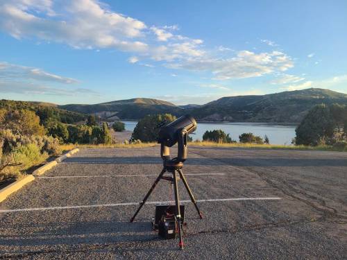 A telescope set up on a parking lot overlooking a serene lake and rolling hills under a clear blue sky.