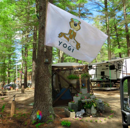 A campsite with a Yogi Bear flag, surrounded by trees, flowers, and a decorative stone arrangement.