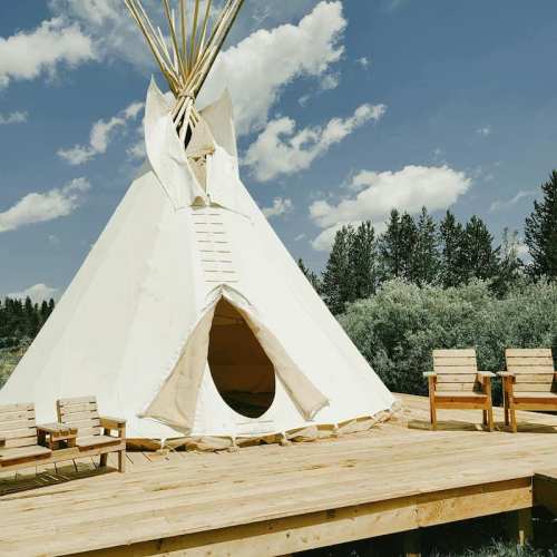 A white teepee on a wooden deck surrounded by trees and a blue sky with fluffy clouds.