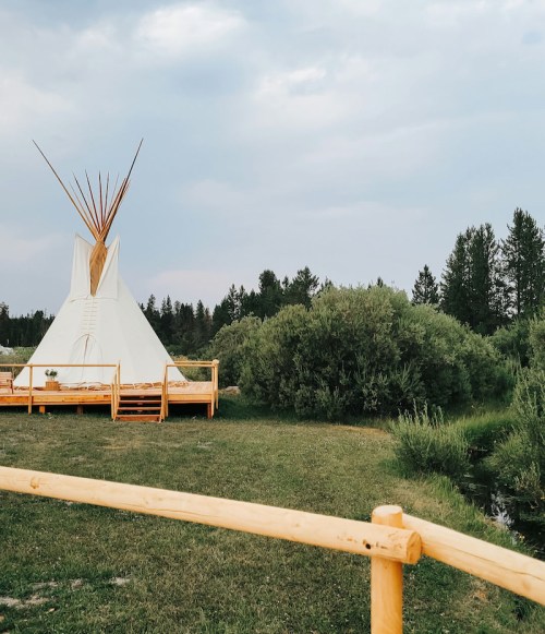 A white teepee stands on a grassy area near a river, surrounded by trees under a cloudy sky.