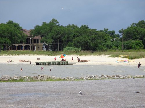 A beach scene with people swimming, an umbrella, and a house in the background, surrounded by trees and a cloudy sky.