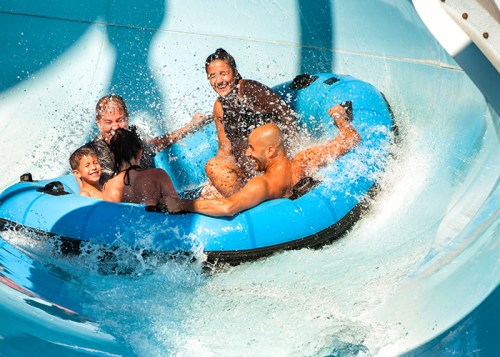 A group of five people enjoying a water slide in a blue inflatable raft, splashing water around them.