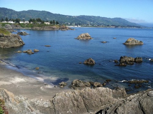 A serene coastal view with rocky shores, calm blue water, and distant hills under a clear sky.