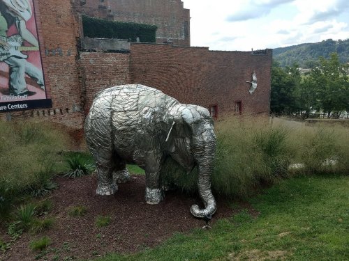 A large silver elephant sculpture stands among greenery near a brick building.
