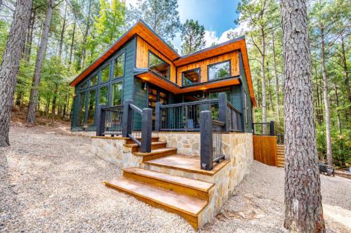 Modern cabin with large windows, wooden accents, and stone steps, surrounded by tall trees and gravel pathway.