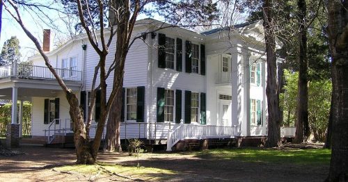 A large white house with green shutters, surrounded by trees and a gravel path, on a sunny day.