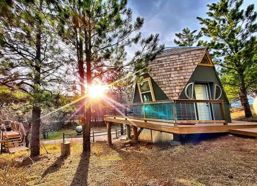 A cozy A-frame cabin surrounded by pine trees, with sunlight shining through, creating a warm, inviting atmosphere.