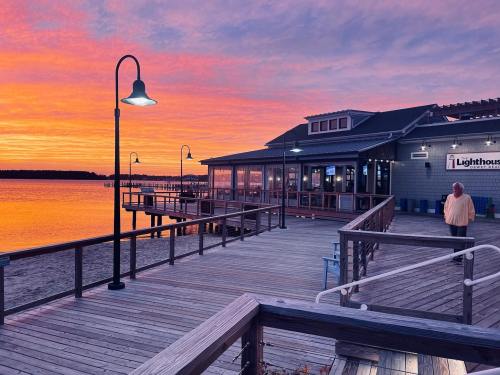 A vibrant sunset casts colorful hues over a waterfront restaurant and wooden pier, with a person walking nearby.