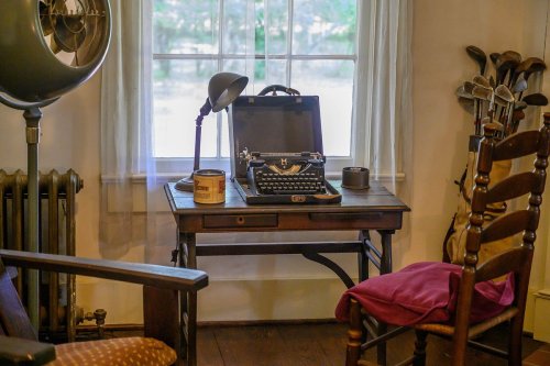 A vintage writing desk with a typewriter, lamp, and chair, set by a window in a cozy room.