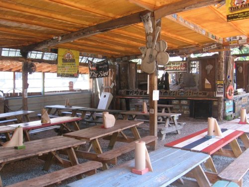 A rustic outdoor dining area with wooden picnic tables, colorful table tops, and a shaded roof, decorated with signs.