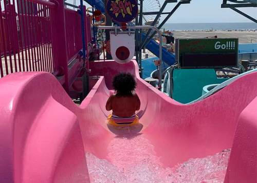 A child with curly hair slides down a pink water slide, splashing into a pool below on a sunny day.