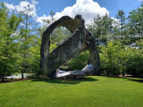 A large abstract sculpture made of stone sits on green grass, surrounded by trees and under a blue sky with clouds.