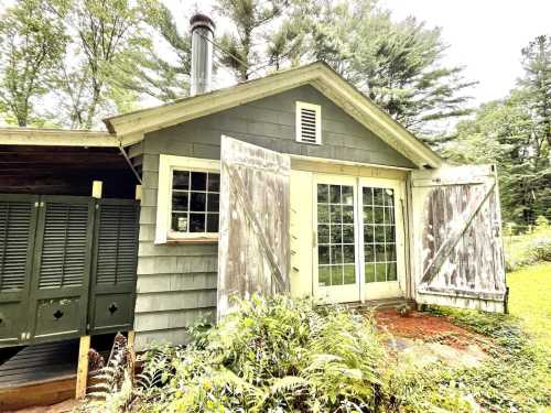 A rustic cabin with weathered shutters, large windows, and a chimney, surrounded by greenery.