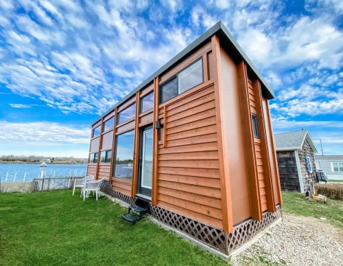A modern tiny house with wooden siding, large windows, and a grassy area, overlooking a body of water under a blue sky.