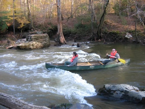 Two people in a canoe navigate a rocky river surrounded by autumn trees.