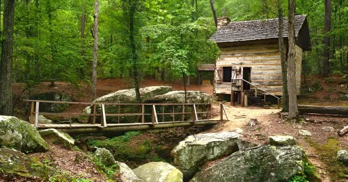 A rustic wooden cabin surrounded by lush green trees and large rocks, with a small bridge crossing a stream.