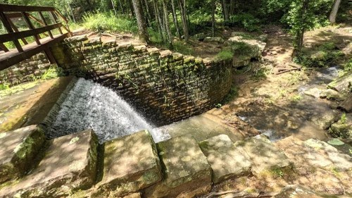 A stone dam with flowing water, surrounded by lush greenery and trees, under dappled sunlight.