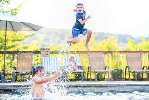 A boy jumps joyfully into a pool while a man splashes below; lounge chairs and greenery are in the background.