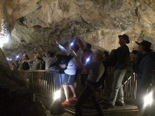 A group of people inside a cave, using flashlights and pointing, with rocky walls and dim lighting around them.