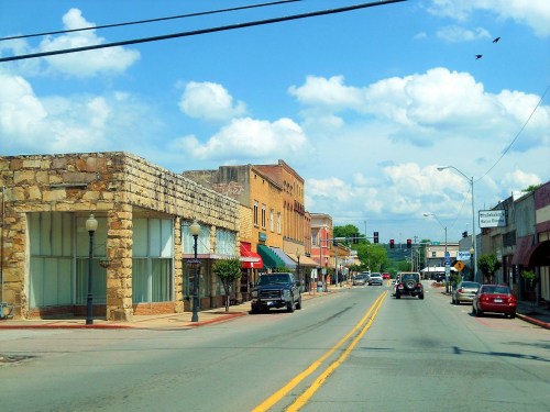 A small town street lined with shops and cars under a blue sky with fluffy clouds.