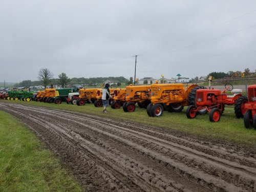 A row of vintage tractors in various colors parked on a muddy field under a cloudy sky.