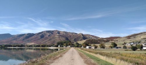 A dirt path leads towards mountains, with a calm lake on one side and fields and houses on the other under a clear sky.