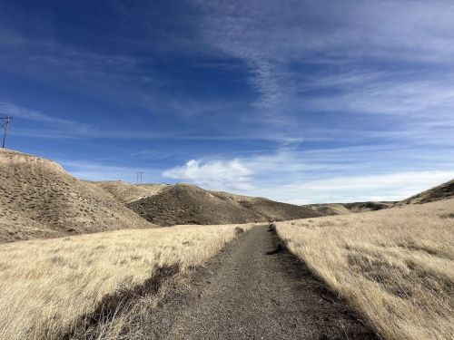 A dirt path winds through dry, grassy hills under a blue sky with scattered clouds. Power lines stretch in the distance.