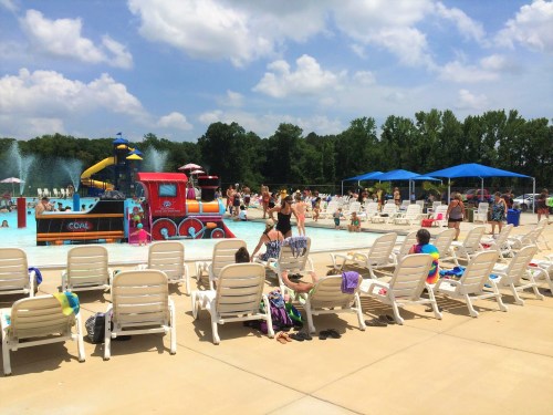 A busy outdoor pool area with families enjoying the water, colorful slides, and lounge chairs under blue skies.