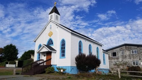 A white church with blue accents and a cross on top, surrounded by greenery and a cloudy sky.
