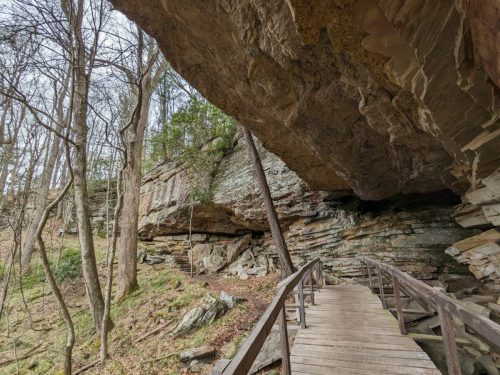 A wooden bridge leads through a rocky area with trees and a large overhanging rock formation above.
