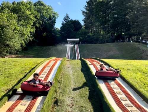 Two children sit in inflatable tubes at the top of a grassy slide, ready to go down on a sunny day.