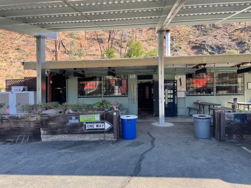 A small roadside store with outdoor seating, surrounded by trees and hills, featuring signs and a trash bin.