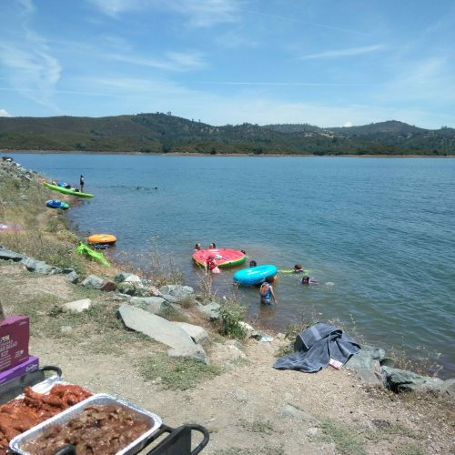 A lakeside scene with people swimming and floating on colorful inflatables, while food is being grilled nearby.