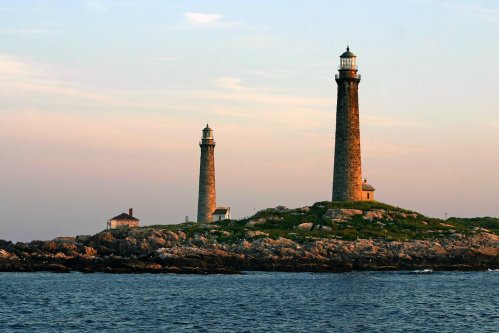 Two lighthouses stand on a rocky island at sunset, surrounded by calm waters and a pastel sky.