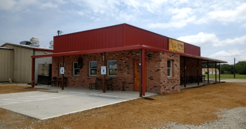 A brick building with a red metal roof, featuring outdoor seating and signage, set against a cloudy sky.