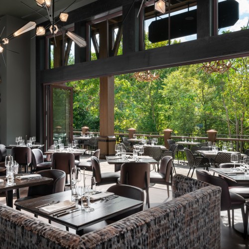 A modern restaurant interior with elegant tables and chairs, large windows overlooking a lush green outdoor area.