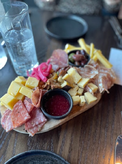A wooden platter with assorted cheeses, cured meats, olives, pickled onions, and a small bowl of sauce, alongside a glass of water.