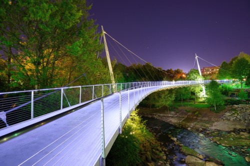 A illuminated suspension bridge at night, surrounded by trees and a calm stream, under a starry sky.