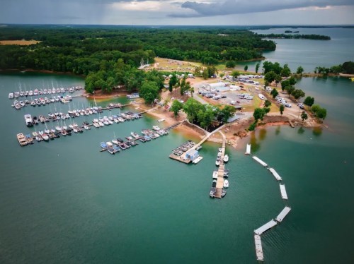 Aerial view of a marina with boats docked, surrounded by green trees and water under a cloudy sky.
