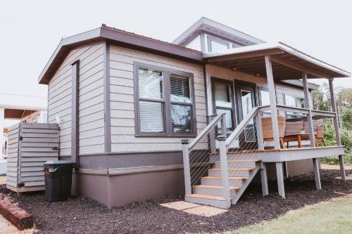 A modern, single-story house with a porch, steps, and large windows, surrounded by mulch and a trash bin.