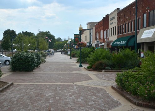 A brick-paved street lined with shops and greenery, under a cloudy sky. Flags and planters add color to the scene.