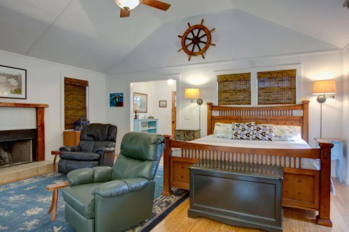 Cozy bedroom with a wooden bed, armchairs, a fireplace, and nautical decor, featuring a ship's wheel on the wall.