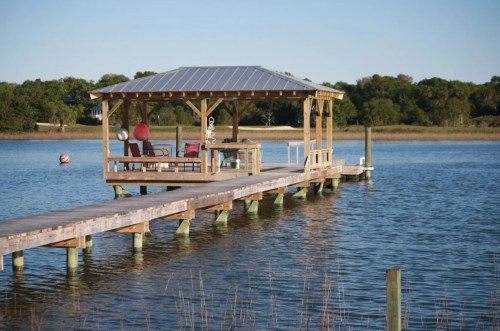 A wooden dock with a gazebo extends over calm water, surrounded by trees and a clear blue sky.