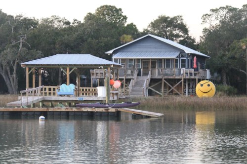 A waterfront house with a dock, kayak, and a large smiley face decoration in the yard, surrounded by tall grass.